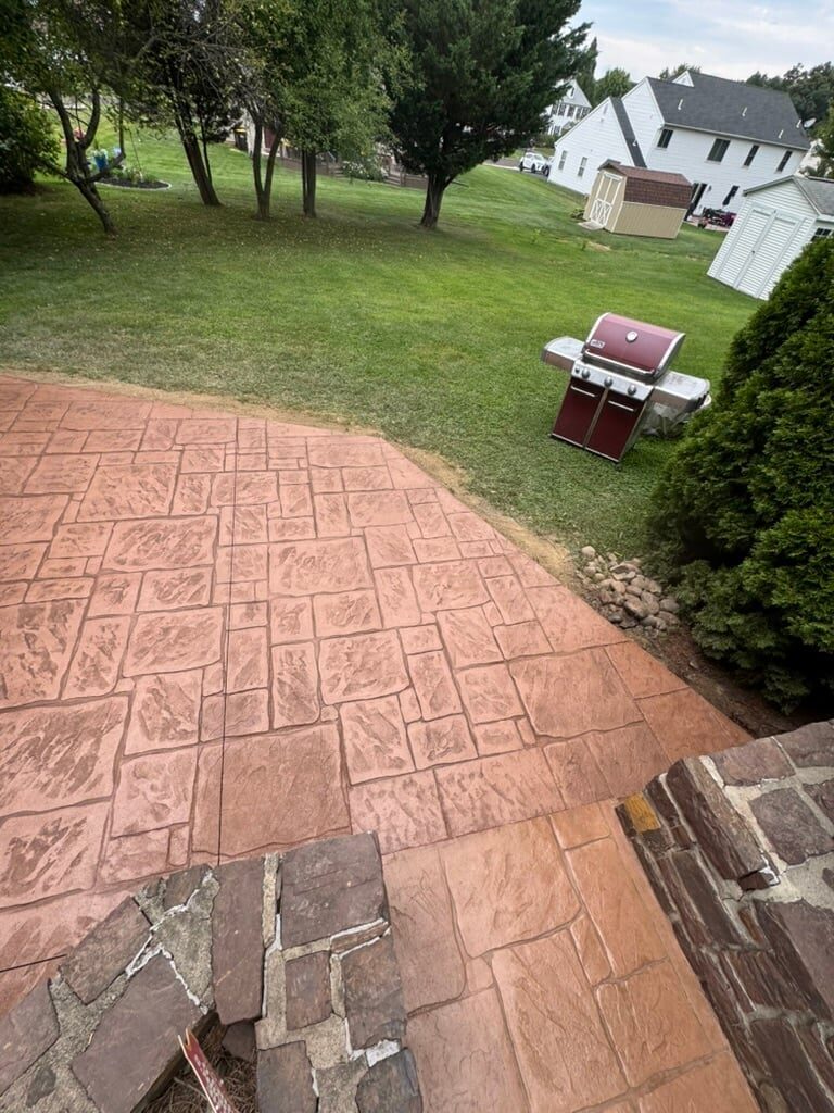 Backyard patio with red brick pavers, gas grill, lush green lawn, shade trees, and white house in background