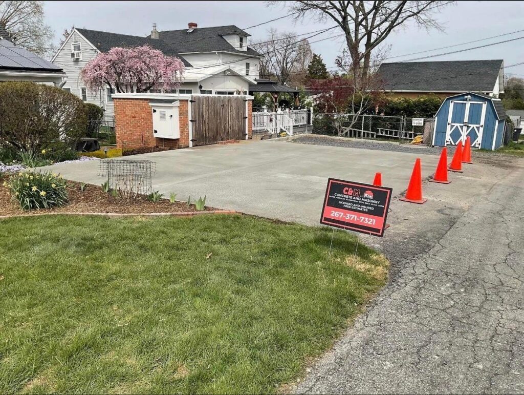 Residential driveway with red traffic cones, real estate sign, storage sheds, and blooming cherry tree in spring